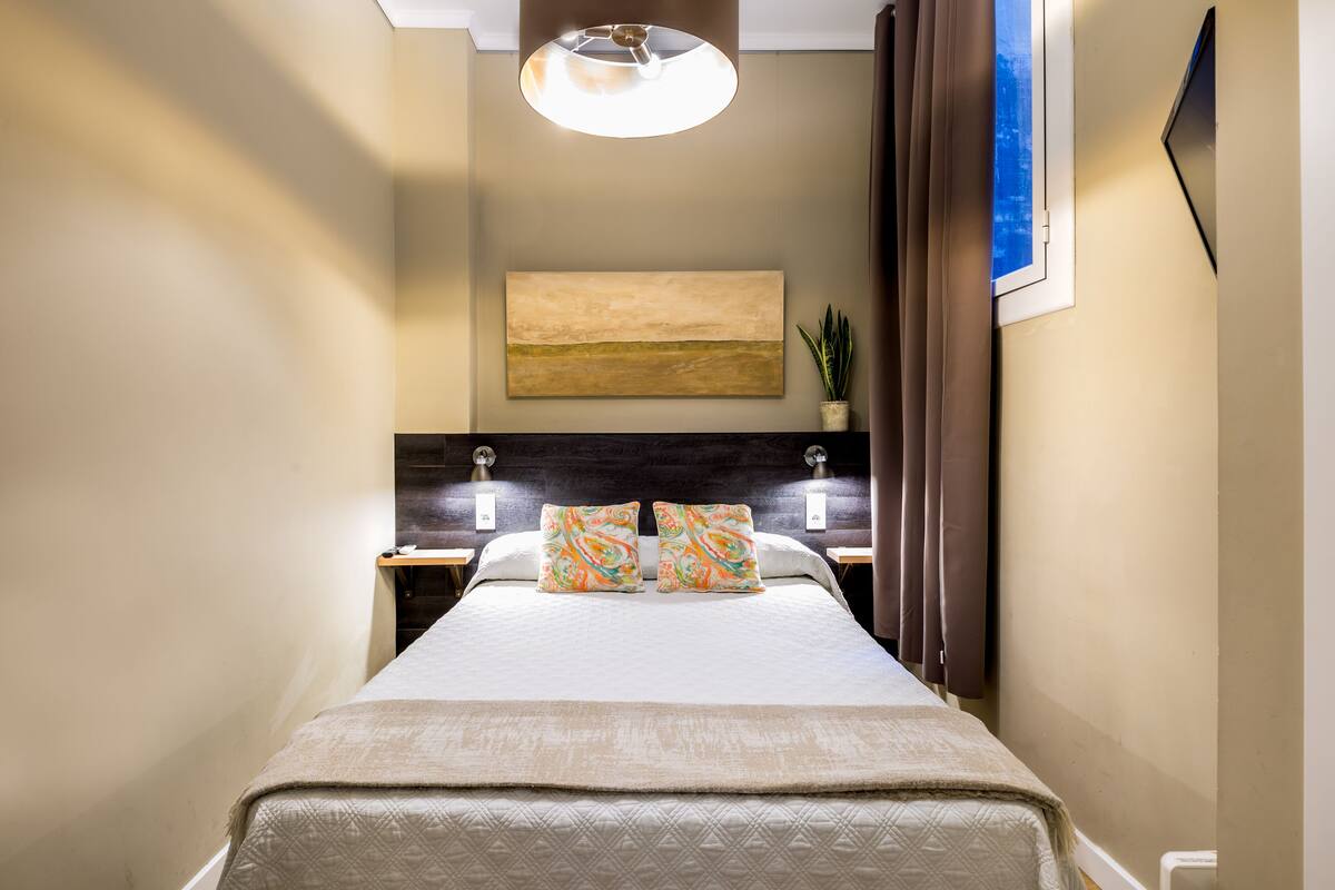 Ein Hotelzimmer mit Bett, Nachttisch, Wandlampe, einem Gemälde, einer Topfpflanze und einem Fenster mit Vorhängen.