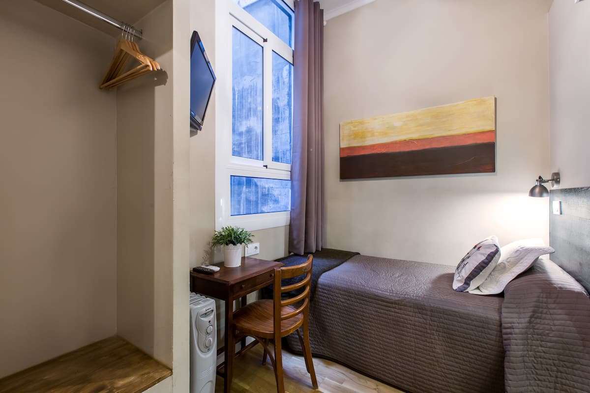 Ein kleiner, gut beleuchteter Raum mit einem Bett, einem Schreibtisch mit Stuhl, einer Topfpflanze und einem fenster mit Vorhängen.