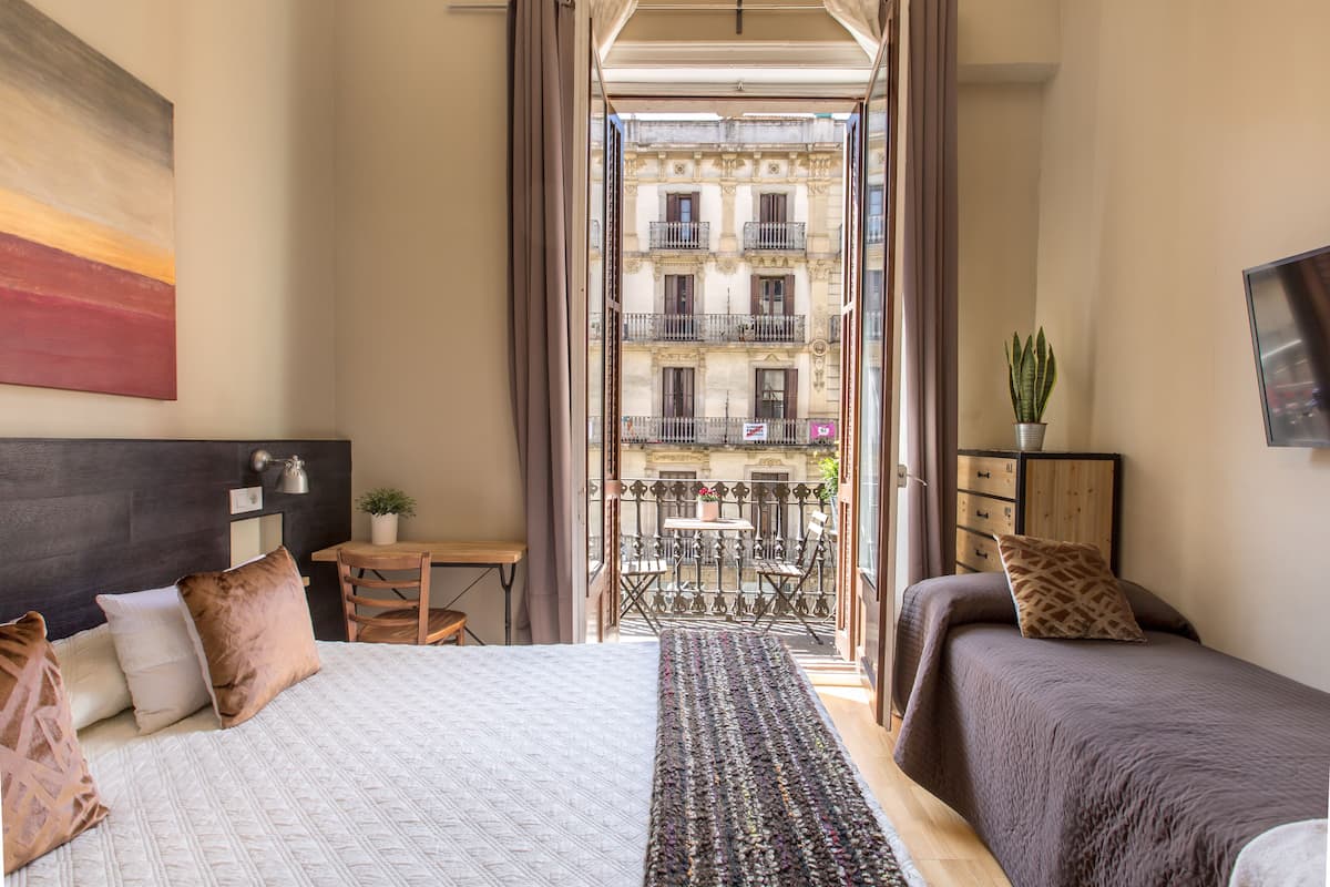 Ein Hotelzimmer mit Bett, Schreibtisch, Stuhl, kleinem Tisch, einer Topfpflanze und einem Balkon mit Blick auf Gebäude.