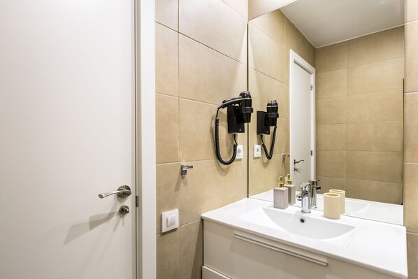 A bathroom with beige tiles, a white sink, two black hair dryers mounted on the wall, and a mirror reflecting the room.