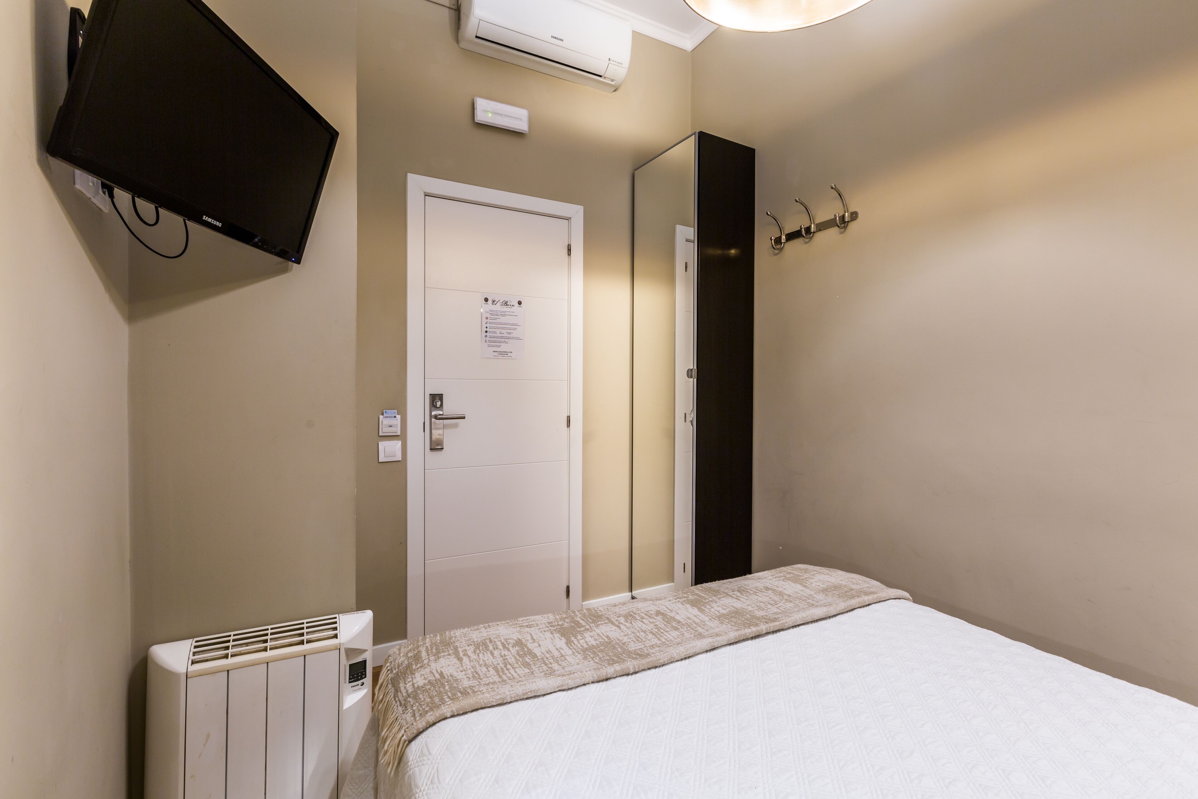Habitación de hotel con cama, televisor montado en la pared, puerta con un letrero, radiador y perchero con ganchos.