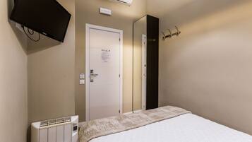 Habitación de hotel con cama, televisor montado en la pared, puerta con un letrero, radiador y perchero con ganchos.