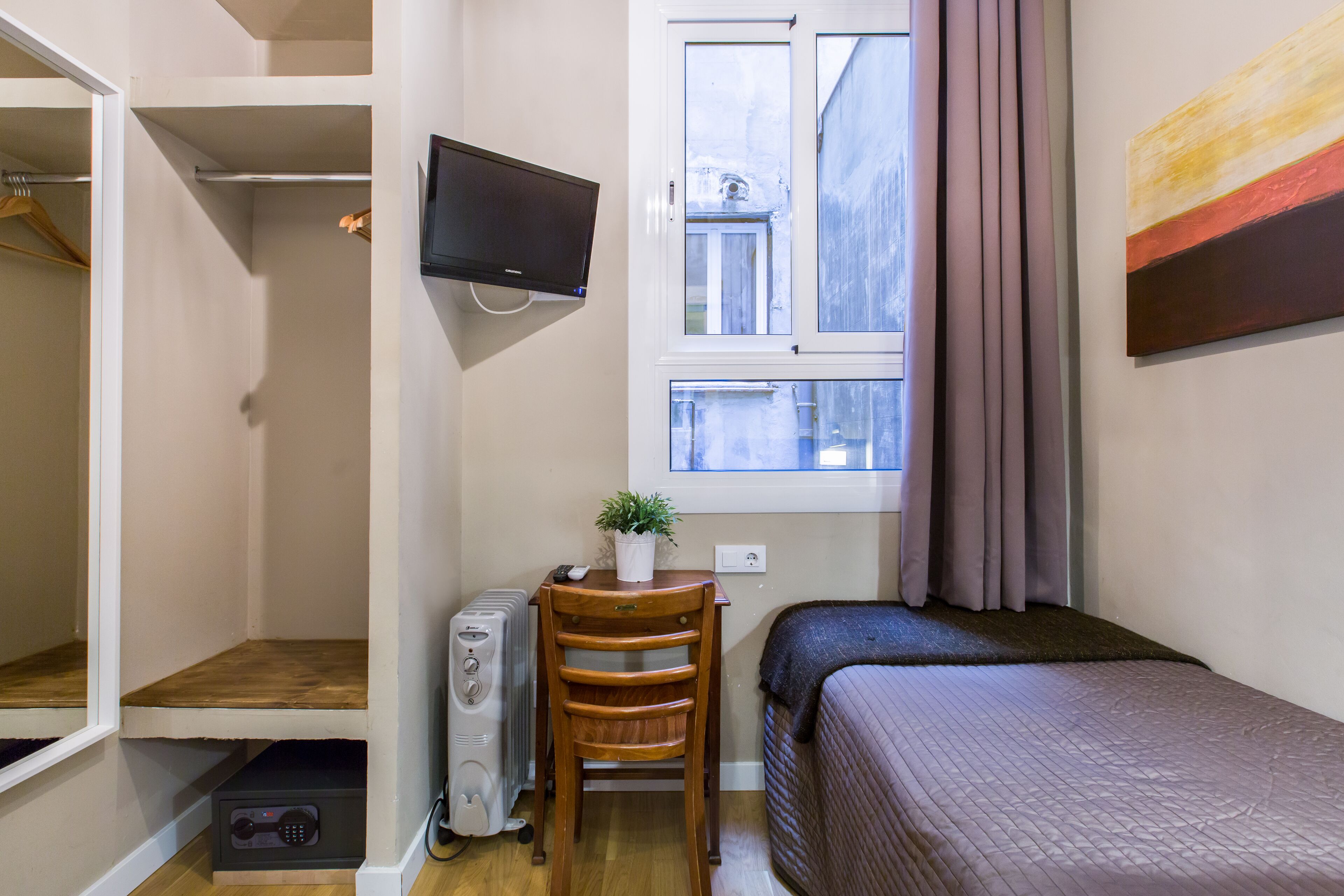 Un dormitorio pequeño con cama, escritorio con ordenador, silla, radiador, televisión y ventana con cortinas.