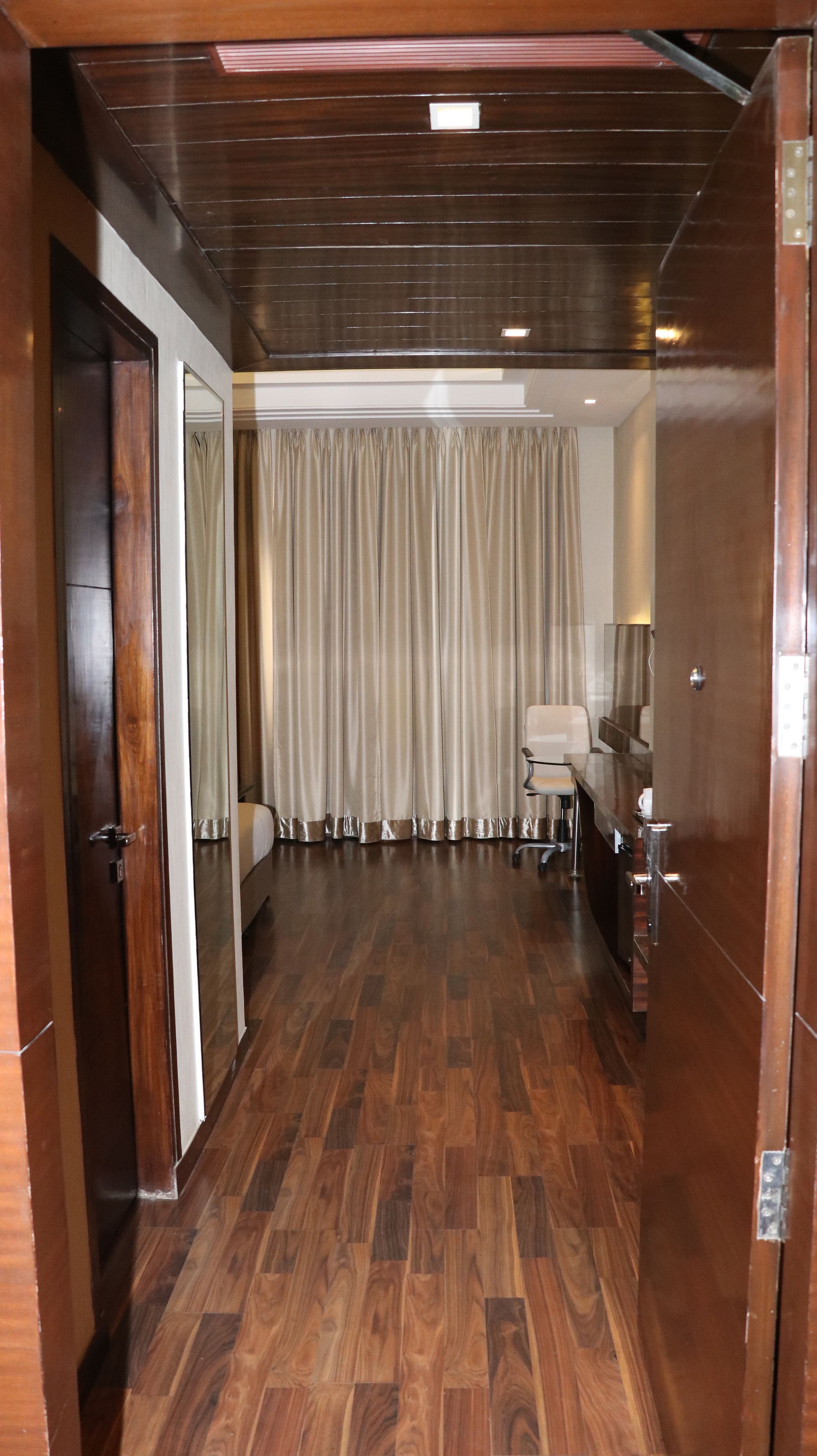 Een hotelkamer met een houten vloer, een bed, een bureau met stoel, een raam met gordijnen, en een deur.