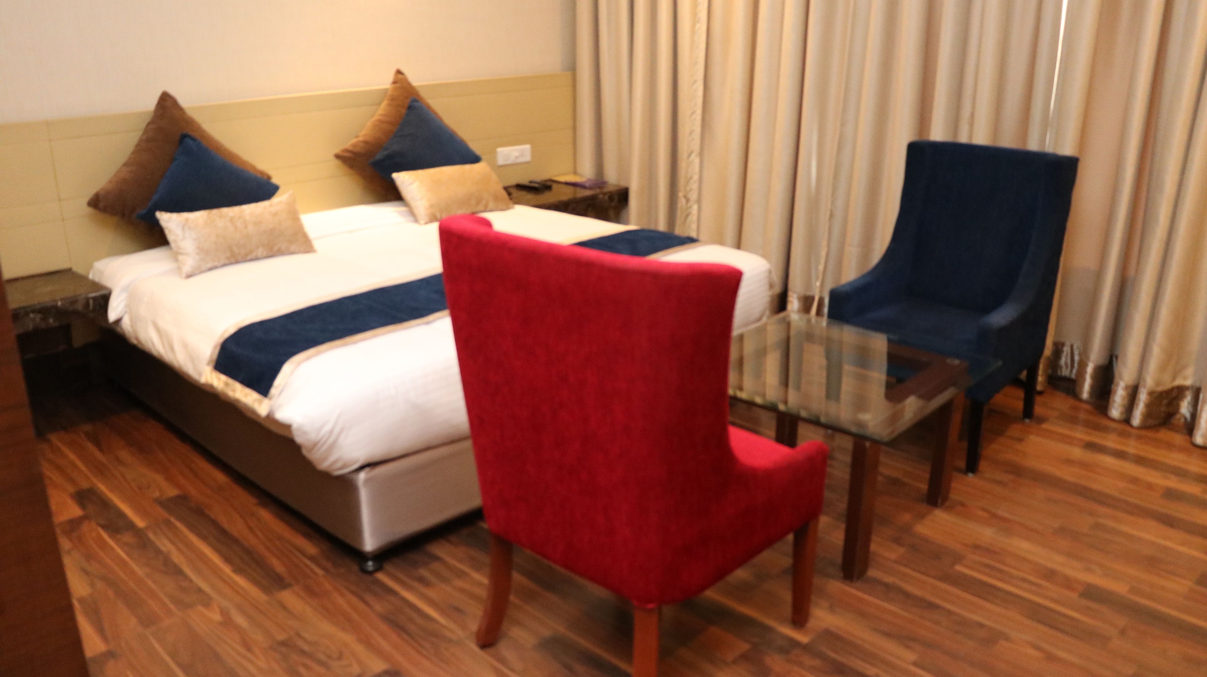 A hotel room with a bed, a glass table, a red chair, and a blue chair.