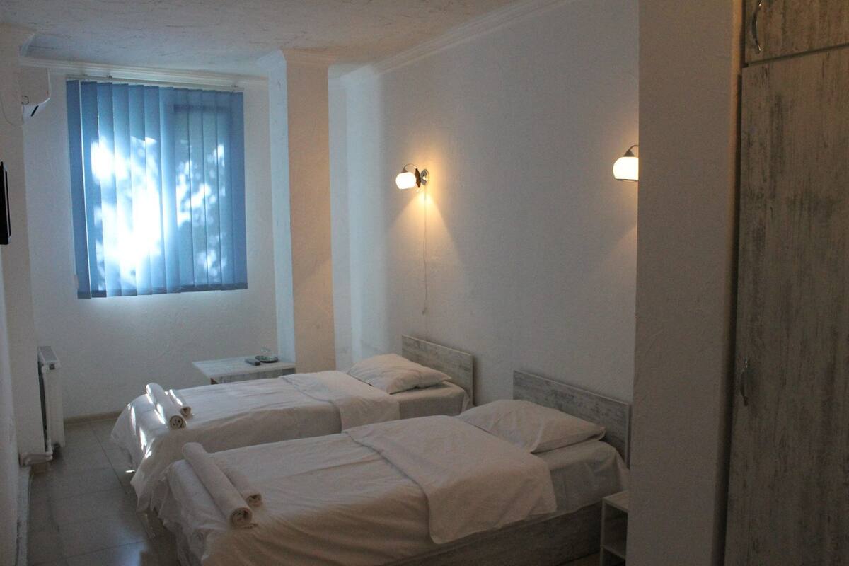 A hotel room with two single beds, a small table, a window with curtains, and wall-mounted lights.