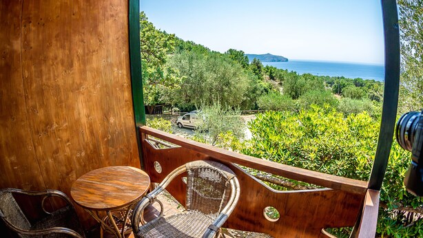 Ein Holzbalkon mit Korbstühlen und einem kleinen Tisch, mit Blick auf eine üppige grüne Landschaft und das Meer.
