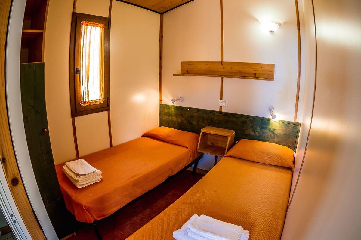 Ein Zimmer mit zwei Einzelbetten, einem kleinen Nachttisch, einem Fenster mit Vorhängen und einem an der Wand befestigten Regal.