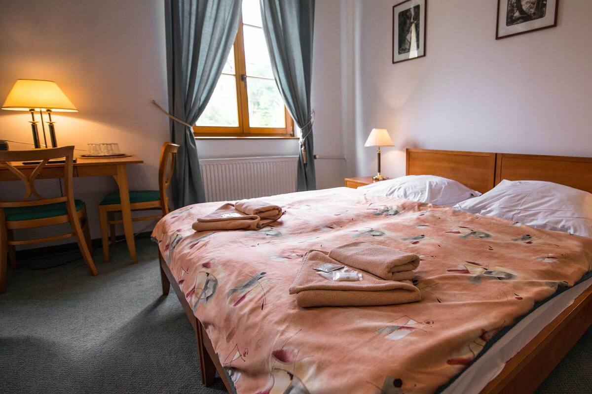 Ein ordentlich bezogenes Bett mit floralem Überwurf und Handtüchern, ein Nachttisch aus Holz mit einer Lampe, ein Sessel und ein Fenster mit Vorhängen.