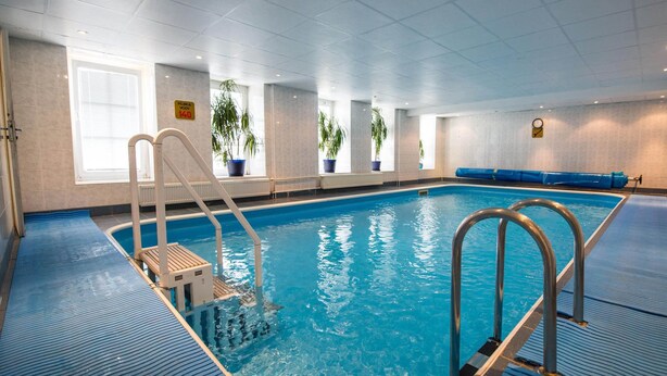 An indoor swimming pool with clear blue water, a diving board, and potted plants along the walls.