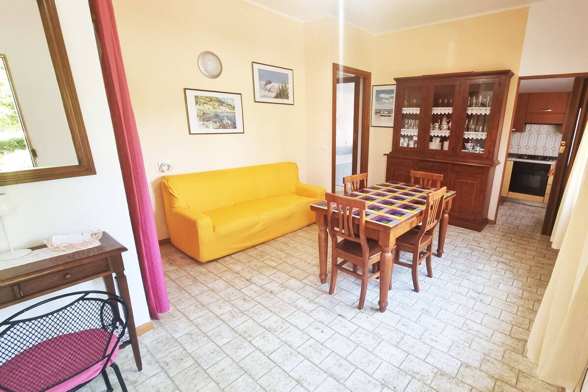 Een woonkamer met een gele bank, een houten eettafel met stoelen, een keuken met kastjes en een betegelde vloer.