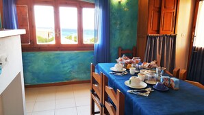 A dining area with a table set for two, a view of the sea through large windows, and a blue curtain. - La Baia Apartments (San Vero Milis)