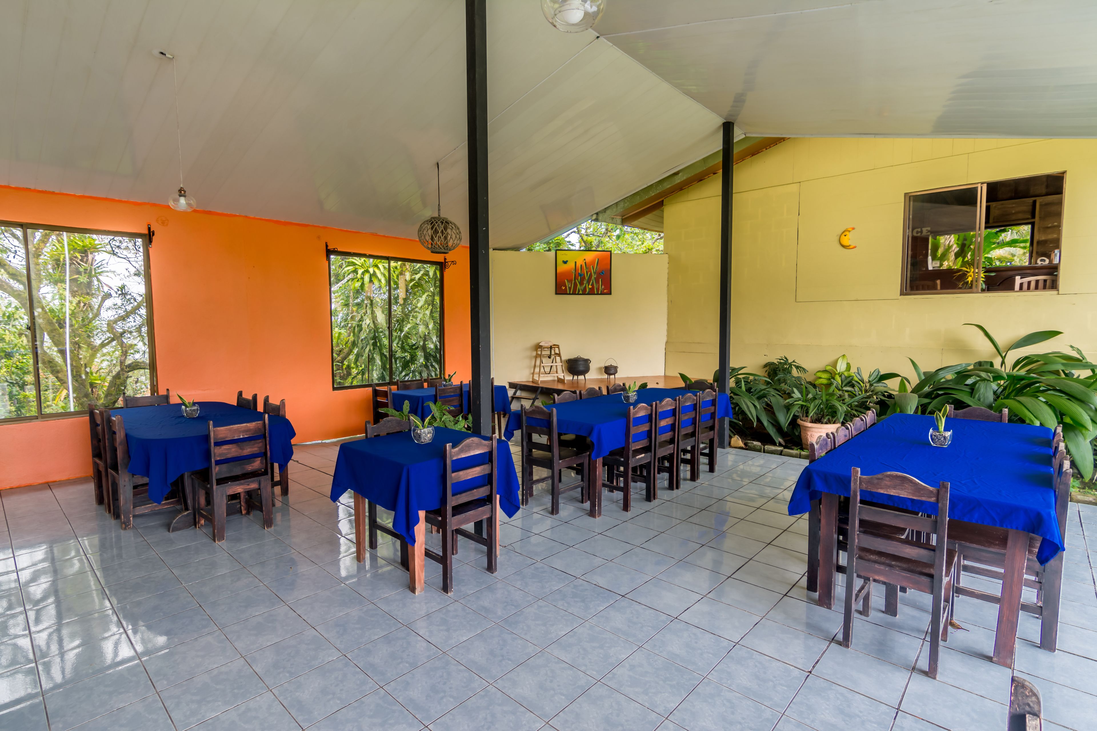 Een restaurant met meerdere tafels, bedekt met blauwe tafelkleden, houten stoelen en plantenbakken met planten.