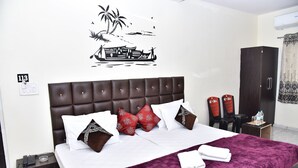 Una cama bien hecha con una colcha morada, almohadas blancas y cojines decorativos. Se observa un aire acondicionado instalado en la pared y una pequeña mesa con una botella de agua y vasos.