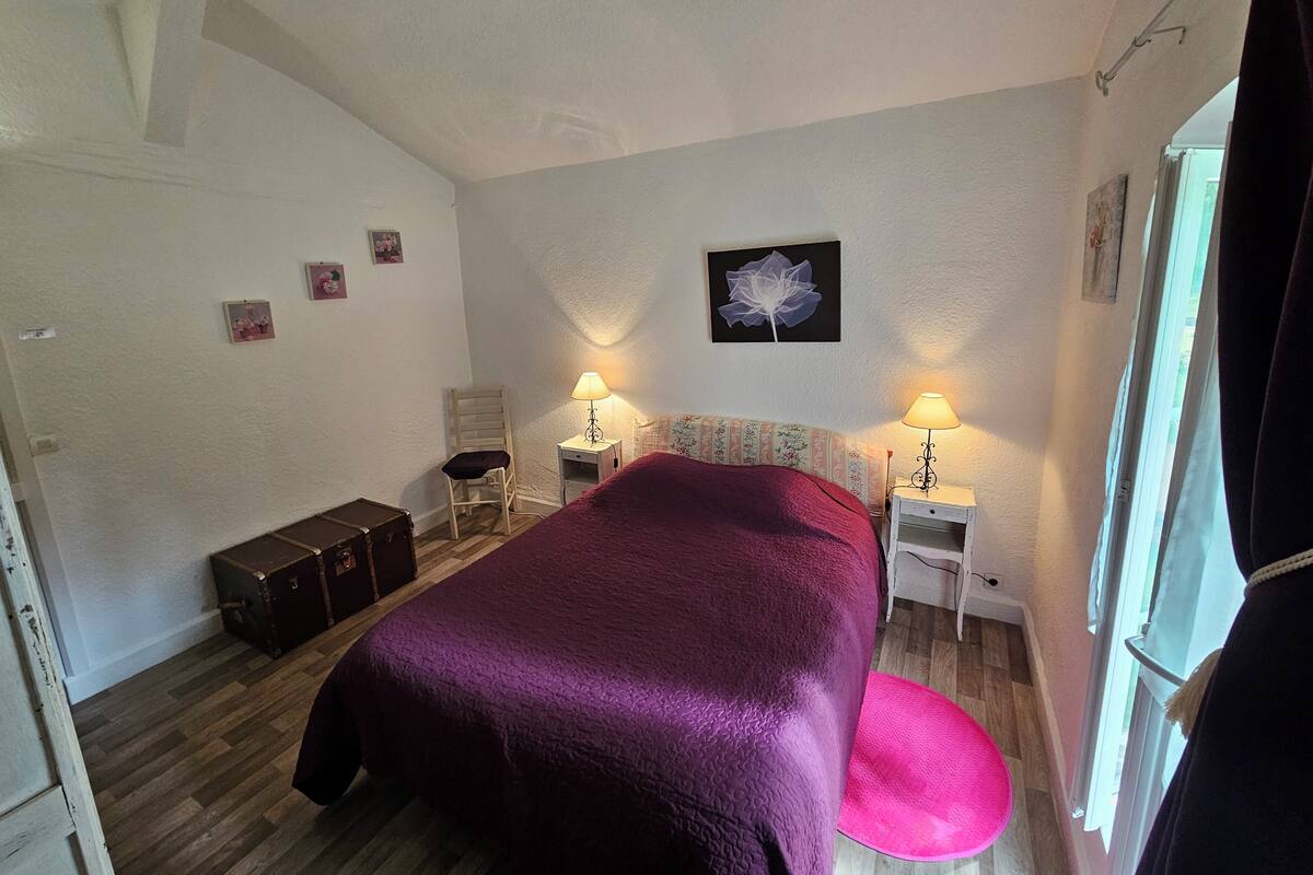 Un dormitorio con una cama grande, dos mesitas de noche con lámparas, una silla, una maleta y una ventana con cortinas.