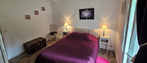 A bedroom with a large bed, two bedside tables with lamps, a chair, a suitcase, and a window with curtains.