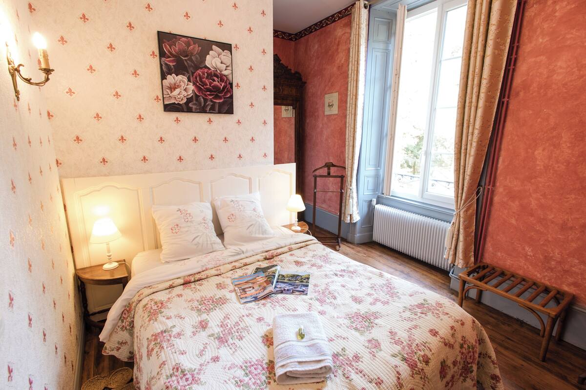 Un dormitorio con colcha estampada, lámpara de noche, cuadro con motivos florales y ventana con cortinas.
