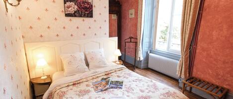 A bedroom with a floral bedspread, a bedside lamp, a framed floral artwork, and a window with curtains.