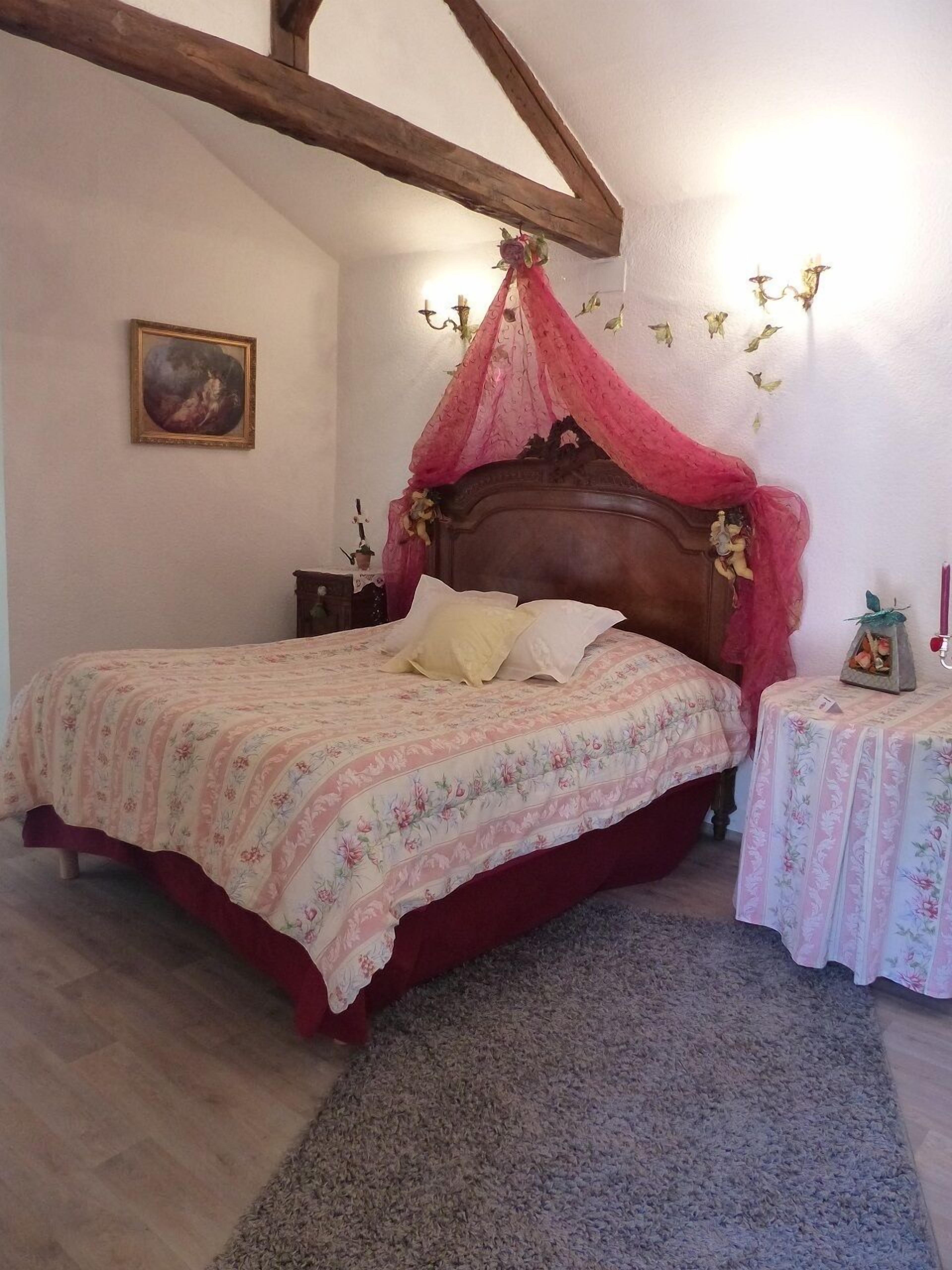 Un dormitorio con cama de madera, ropa de cama floral, dosel, mesita de noche, una pequeña mesa con lámpara y un cuadro enmarcado en la pared.
