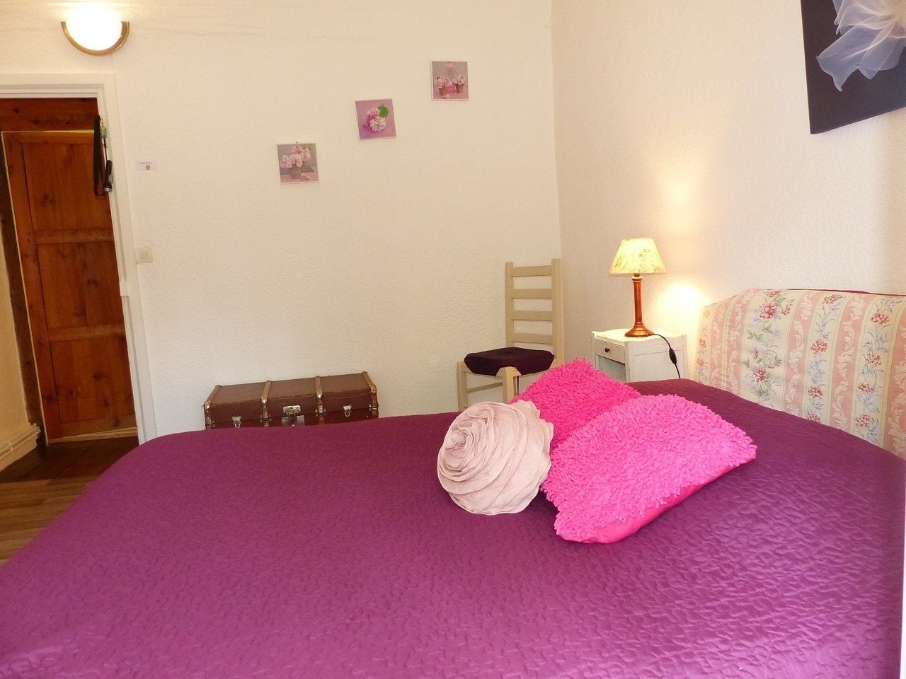 A room with a purple bedspread, a wooden door, a small bedside table with a lamp, and a floral-patterned headboard.