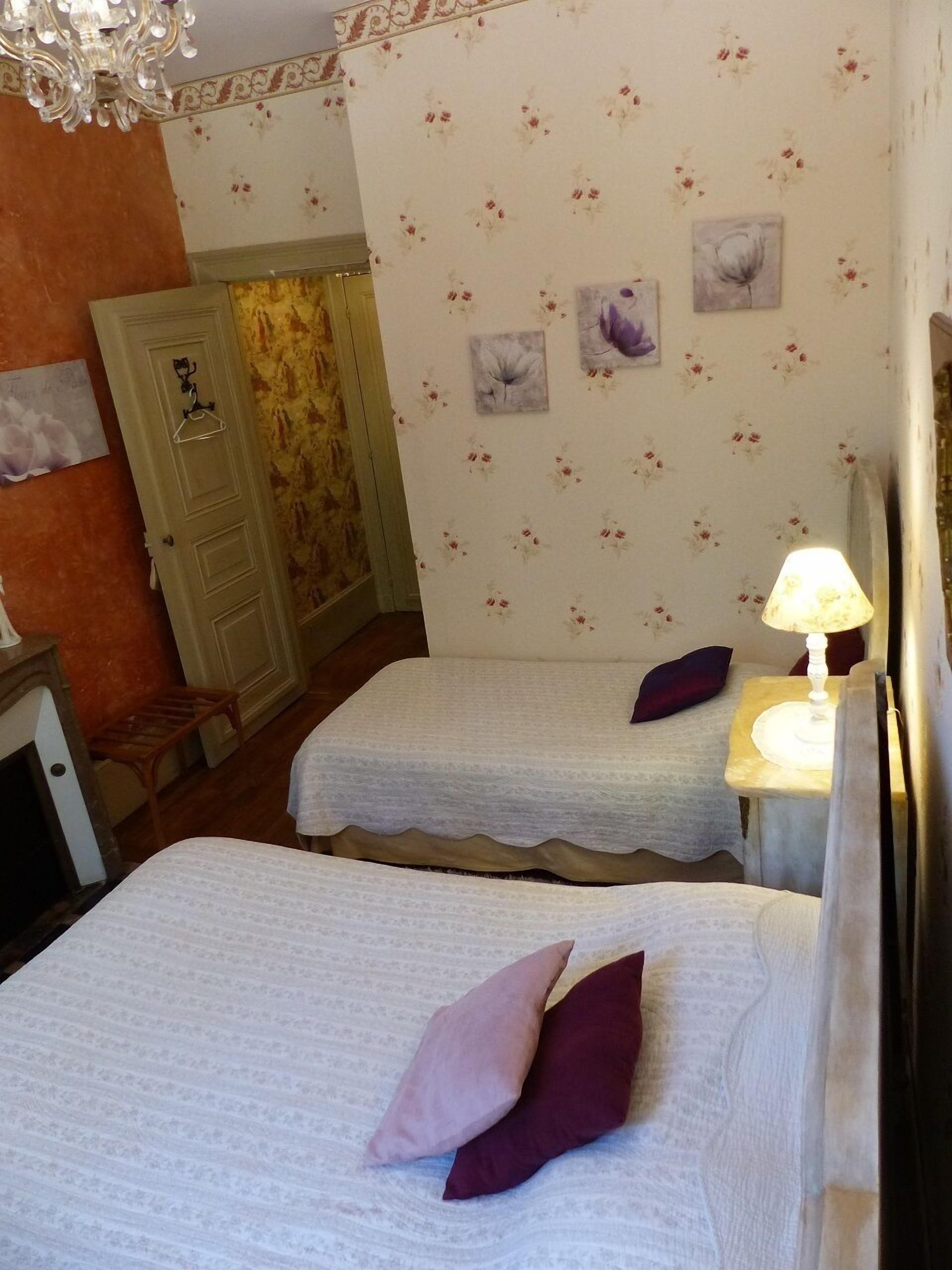 A room with two beds, a small table with a lamp, and floral wallpaper.