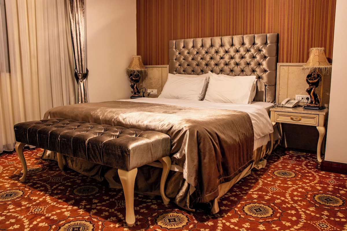 A hotel room with a tufted headboard, a large bed, a bench, a patterned carpet, and a nightstand with a lamp.