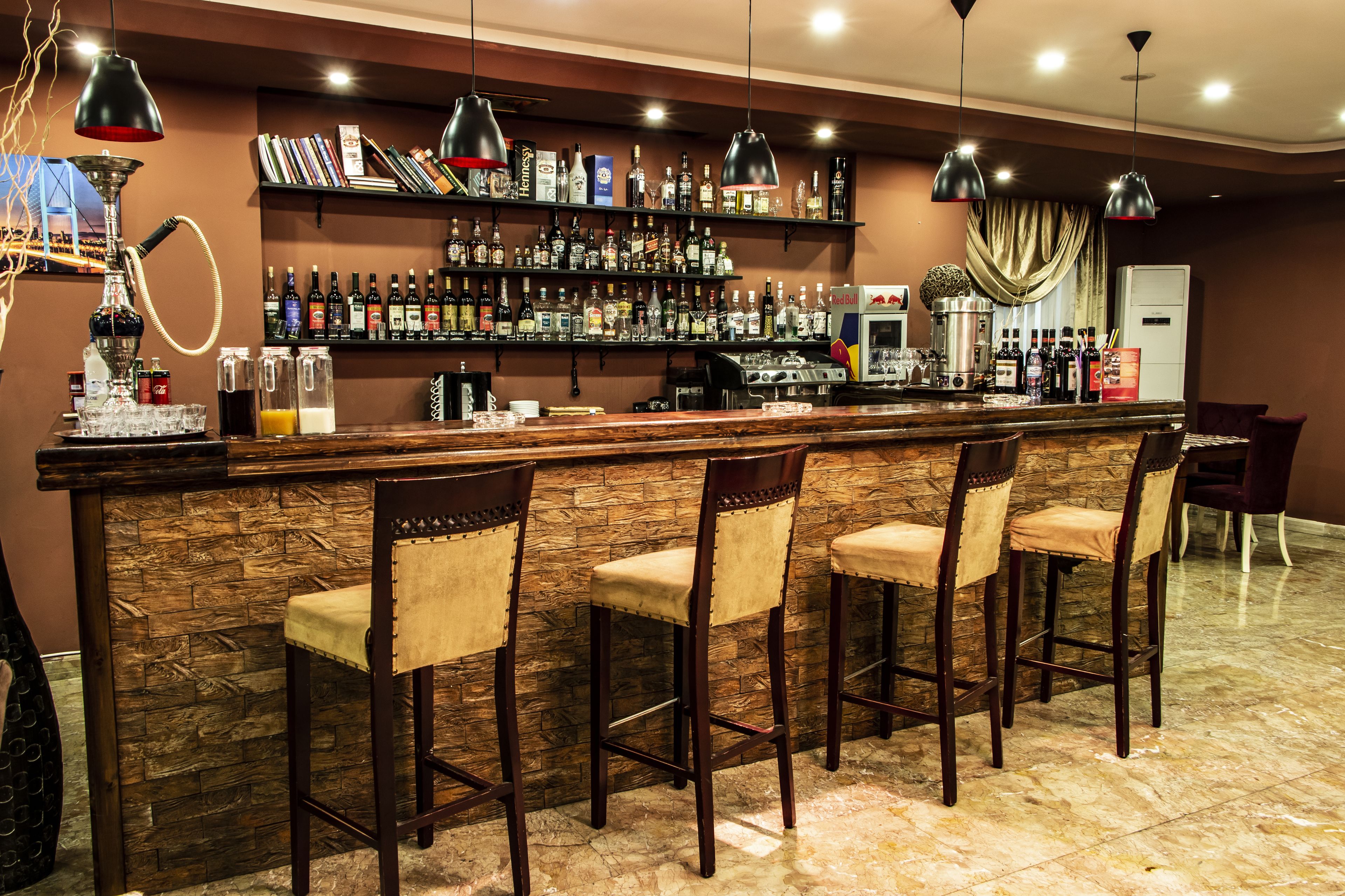 Un'area bar con parete in pietra, bancone in legno e quattro sgabelli da bar. Si notano delle mensole con bottiglie e un narghilè a sinistra.