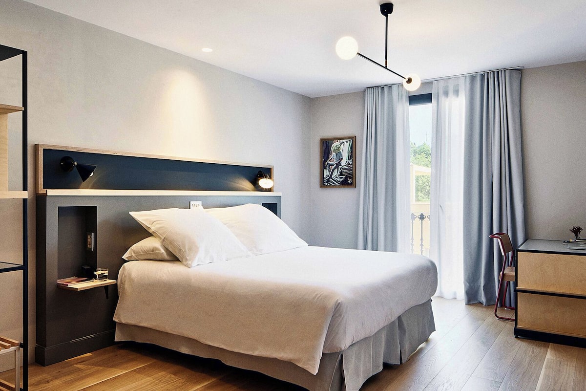 A modern bedroom with a bed, bedside tables, a desk, and a view of the outdoors.