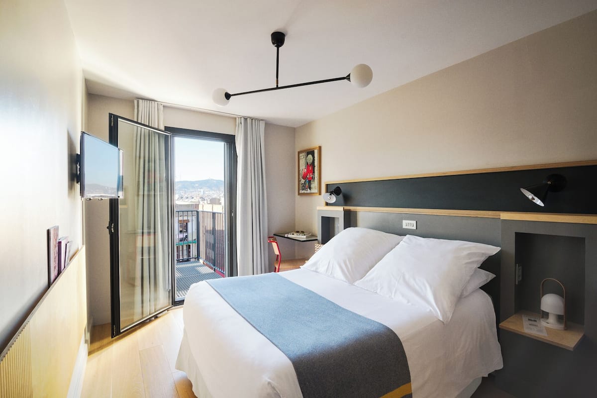 A hotel room with a bed, a desk, a chair, a TV, and a balcony with a city view.