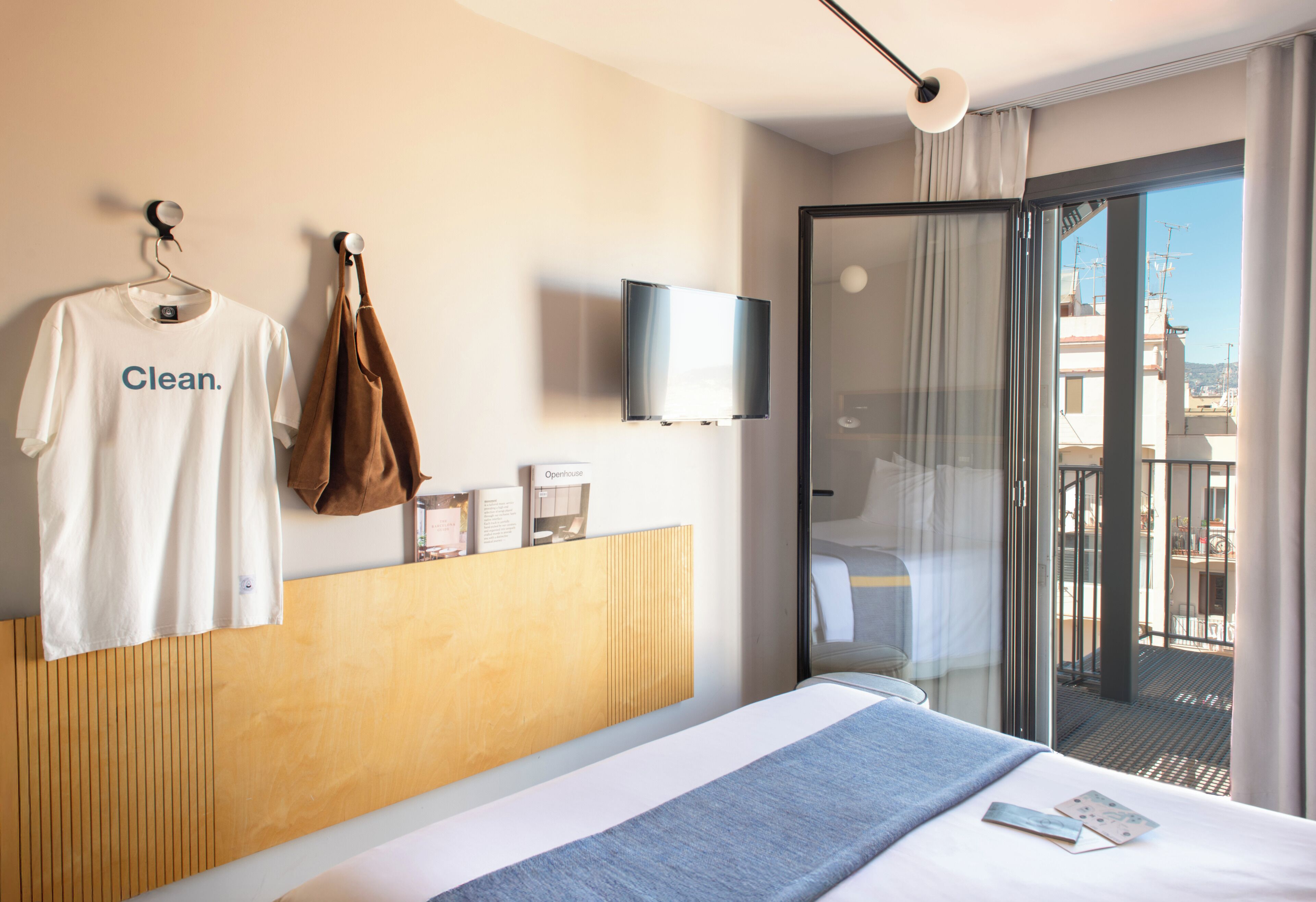Habitación de hotel con cama, televisor montado en la pared, ventana con vista a edificios y abrigo colgado en un perchero.