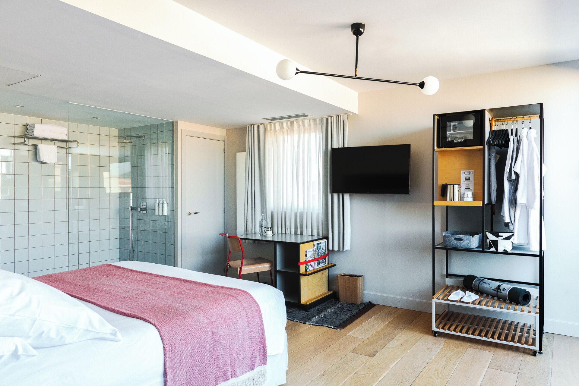 A modern hotel room with a bed, a shower, a desk with a chair, a TV, and a wardrobe.