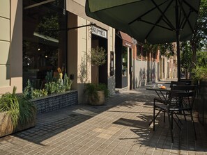 Un área de café al aire libre con mesas, sillas, un cartel que dice 'BRUMMELL' y plantas en macetas.