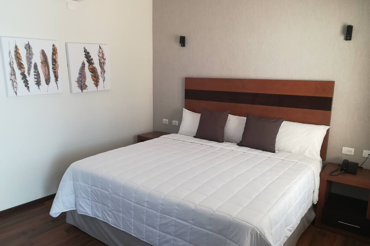 A neatly made bed with a white quilt, a wooden headboard, and a bedside table with a phone.