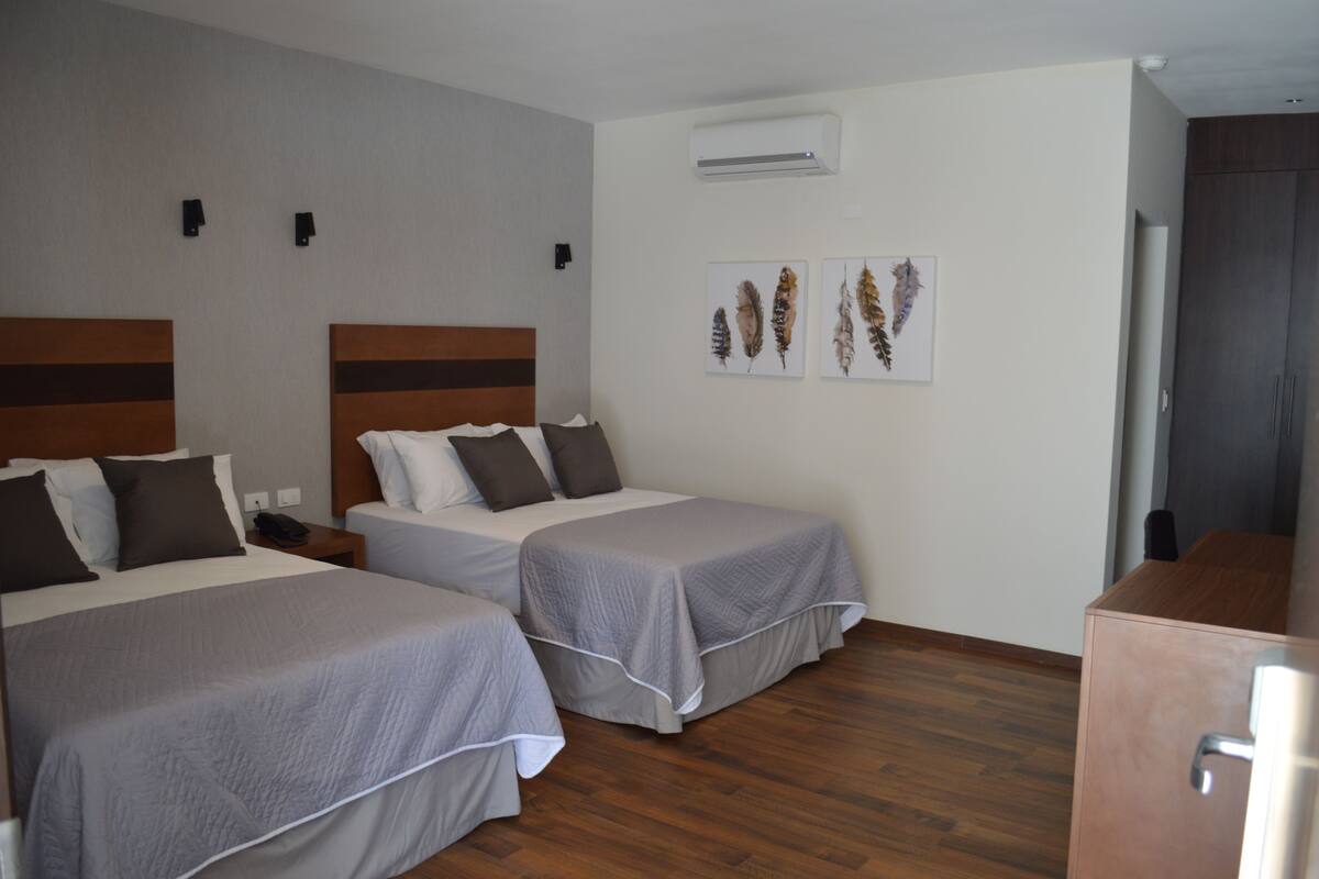 A hotel room with two beds, a wooden floor, an air conditioning unit, and artwork on the wall.