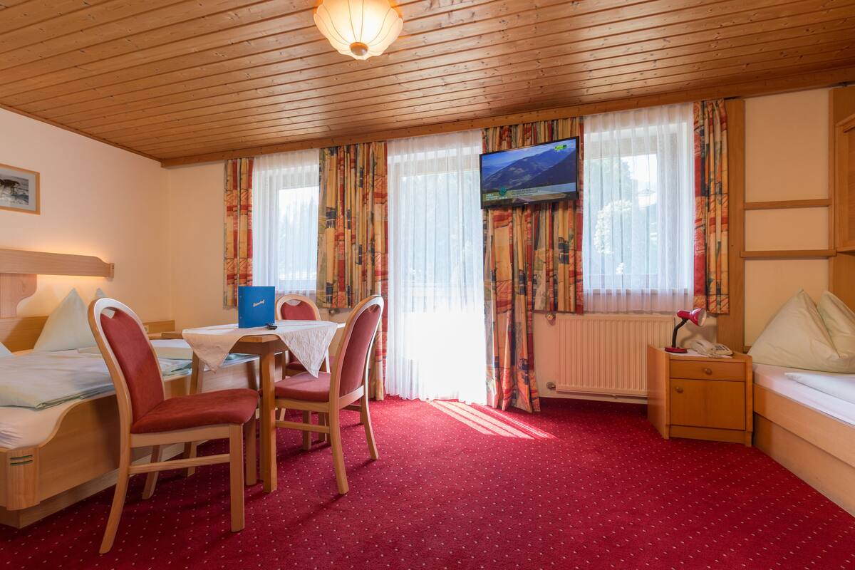 A hotel room with a wooden ceiling, a bed, a desk with a chair, a TV, and a window with curtains.