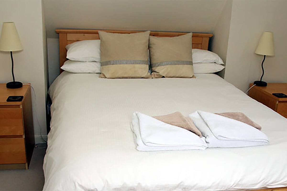 Un lit bien fait, avec du linge de lit blanc et deux oreillers, deux tables de chevet avec des lampes et une tête de lit en bois.