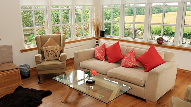 A living room with a beige sofa, red pillows, a glass coffee table, a chair, and a view of greenery through large windows. - Westacre Lodge Self Catering Chalet (Crieff)