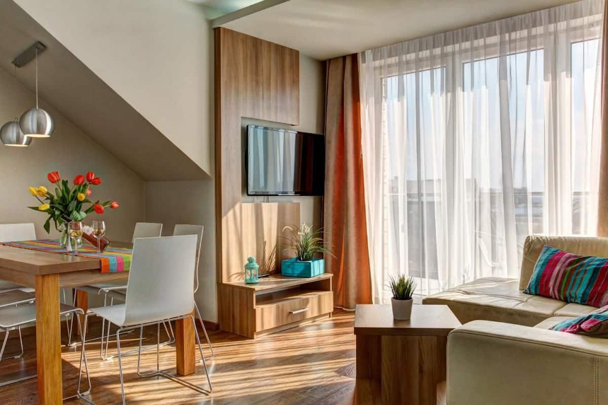 A modern living room with a dining area, wooden flooring, a flat-screen TV, and a large window with curtains.