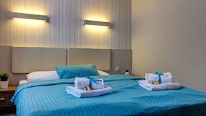 Une chambre d’hôtel avec un grand lit, deux lampes fixées au mur, une table de chevet avec une plante et deux serviettes ornées d’un ruban bleu.