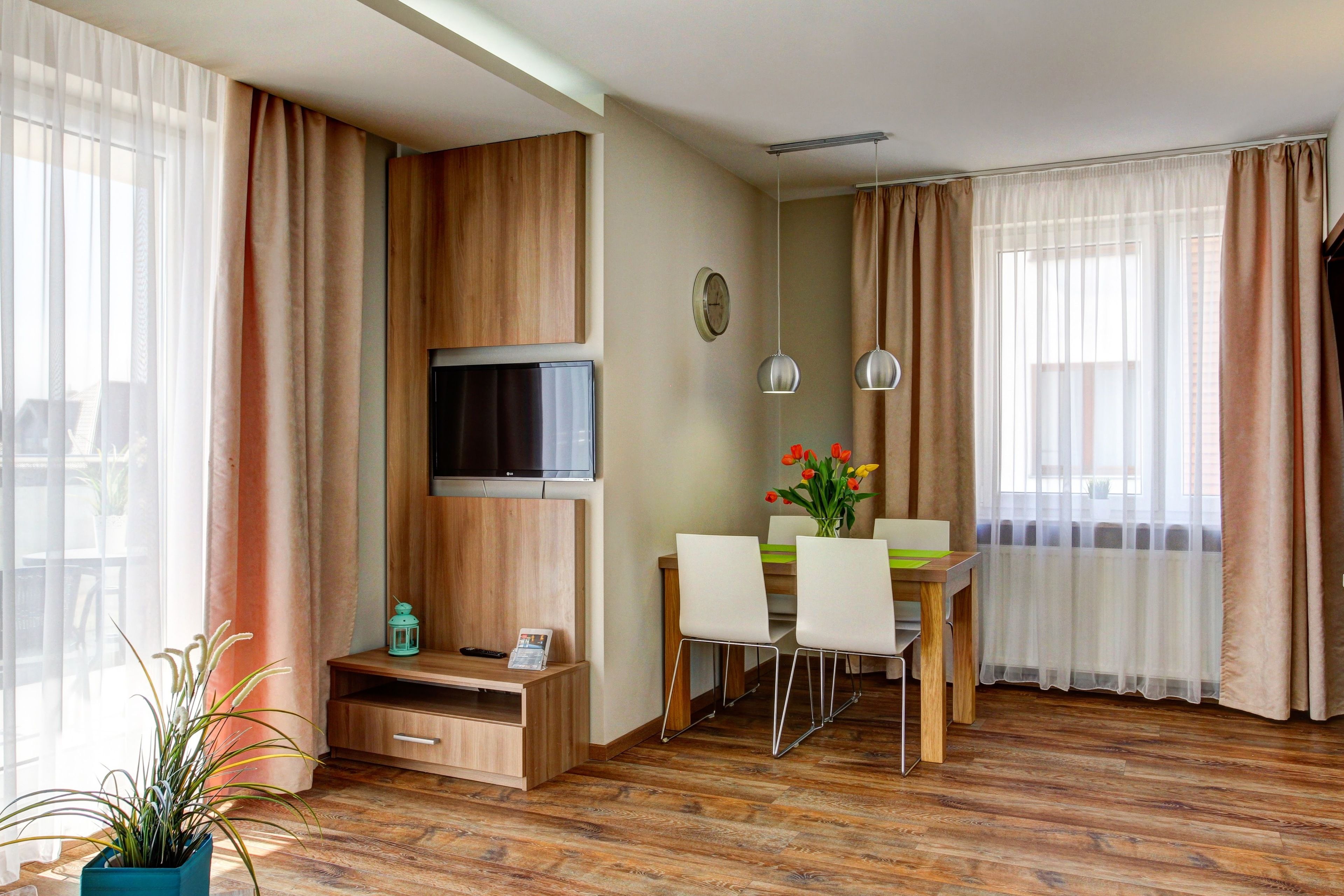 A modern hotel room with a wooden floor, a flat-screen TV mounted on a wooden panel, a dining area with a table and chairs, and large windows with sheer curtains.