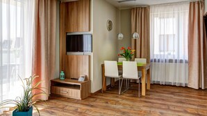 Una habitación de hotel moderna con suelo de madera, un televisor de pantalla plana montado en un panel de madera, una zona de comedor con mesa y sillas, y ventanas amplias con cortinas translúcidas.