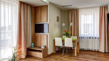 A modern hotel room with a wooden floor, a flat-screen TV mounted on a wooden panel, a dining area with a table and chairs, and large windows with sheer curtains.
