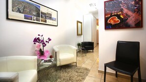 A hotel lobby with a white chair, a black chair, and a glass table with a vase of purple flowers. There are framed pictures on the walls, one showing a cityscape and another showing a building with a circular structure. - Easy Dream Rooms (Verona)