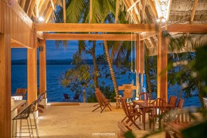 Un espace extérieur aménagé pour les repas, avec du mobilier en bois et une vue sur l’océan au crépuscule.
