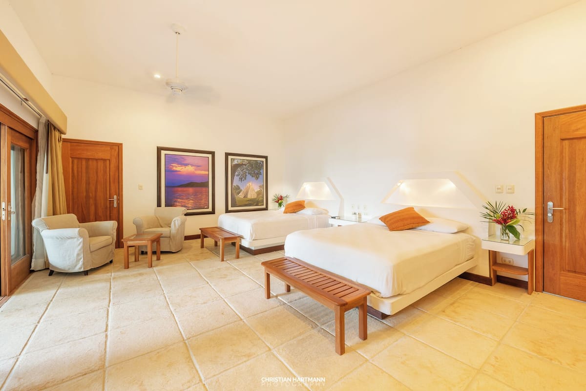 Una habitación de hotel con cama, dos sillas, una mesita, una mesita de noche y una lámpara.