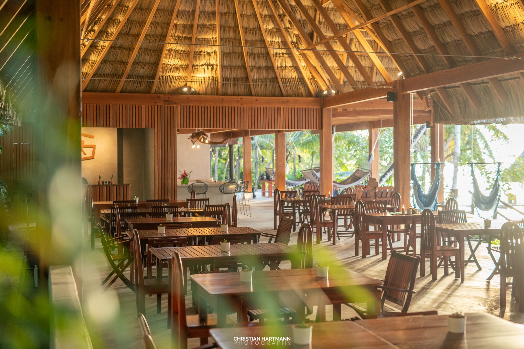 Un espace de restauration en plein air avec des tables et des chaises en bois, un toit de chaume et de grandes fenêtres.