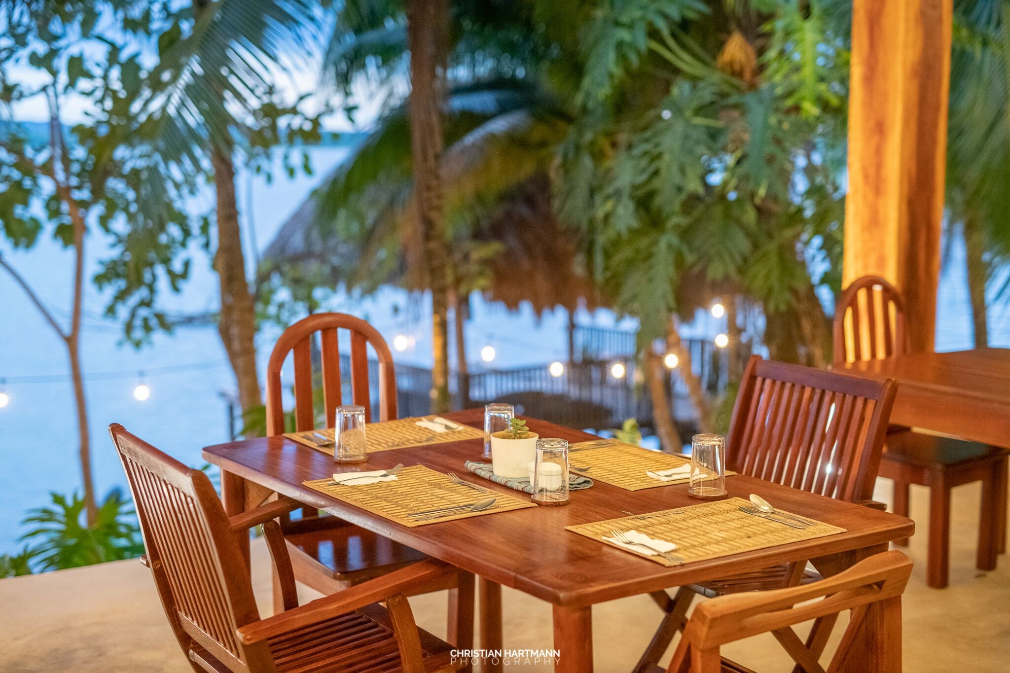 Ein Holztisch gedeckt mit Tischsets, Besteck und Gläsern, umgeben von Stühlen, mit Blick auf Palmen und einen Strand im Hintergrund.