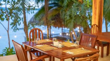 Ein Holztisch gedeckt mit Tischsets, Besteck und Gläsern, umgeben von Stühlen, mit Blick auf Palmen und einen Strand im Hintergrund.