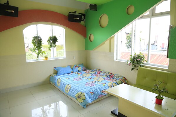 A room with a bed, a small table, and a window with plants.