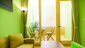 A room with a green wall, a beige sofa, a wooden table, two wooden chairs, a floor lamp, and a sliding glass door leading to an outdoor area with a wooden deck. - Summer Sea Hostel (Magong)